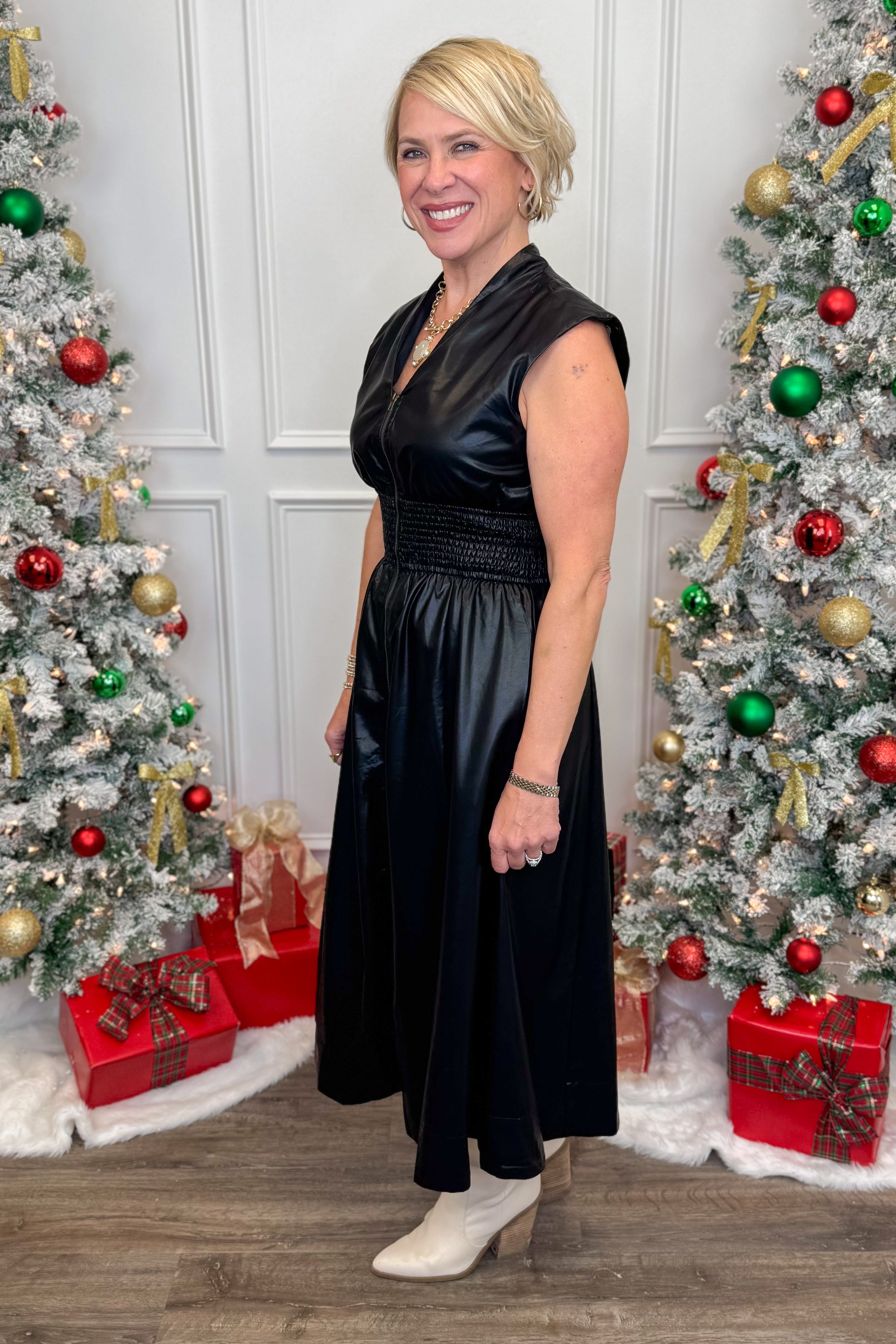 Woman in a black dress standing between two decorated Christmas trees with presents underneath.