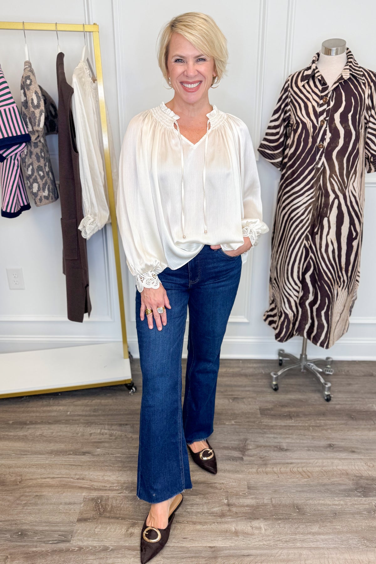Front view of woman wearing Swagger Boutique’s Ivory Satin Cuffed Lace Sleeve Top with jeans with clothing hanging in the background.