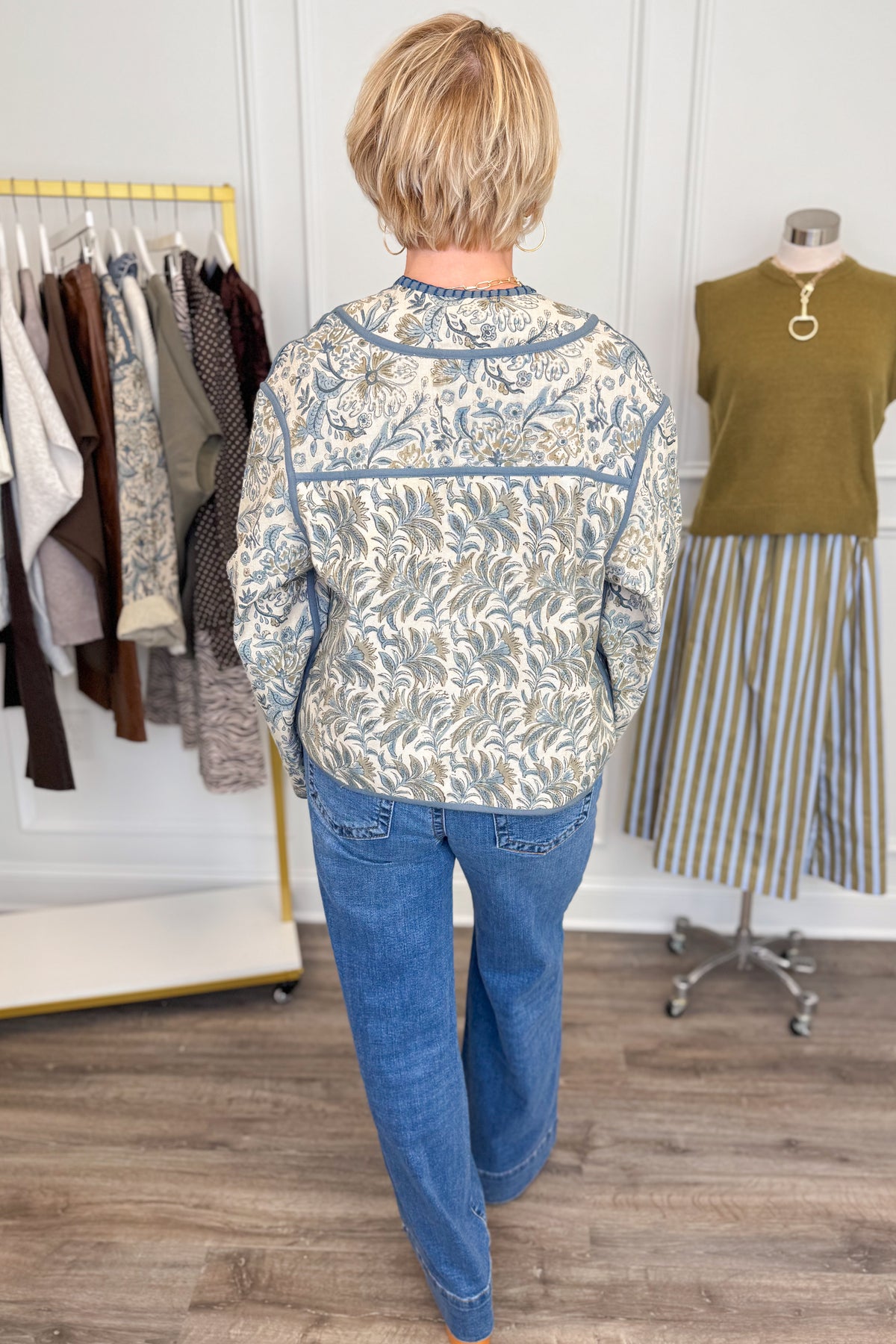 Back view of woman wearing Swagger Boutique’s Beige &amp; Blue Printed Lightweight Jacket, Cream Sleeveless Button up Sweater with Maya Denim Jeans with clothing hanging in the background.