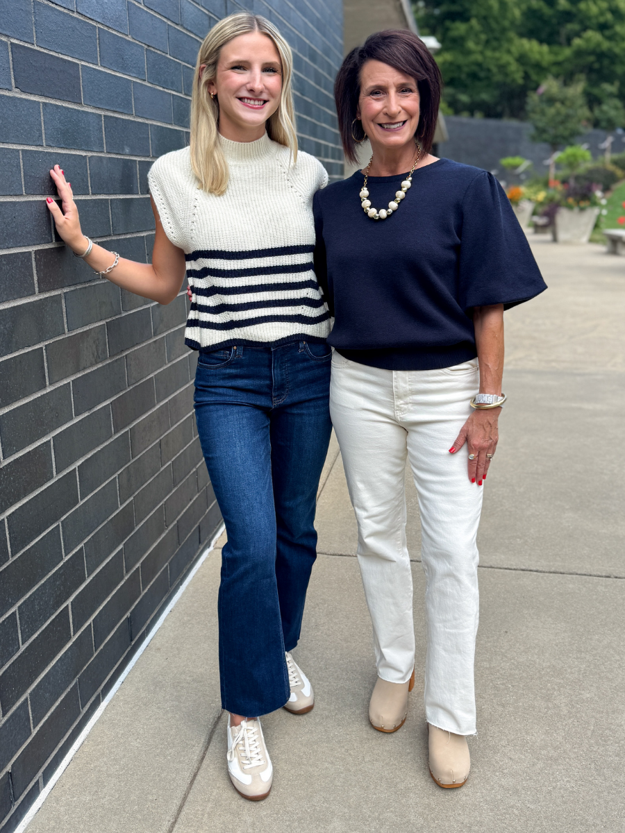 Two women standing outdoors next to a dark gray brick wall. The woman on the left wears a cream and navy striped knit top, blue jeans, and white sneakers.