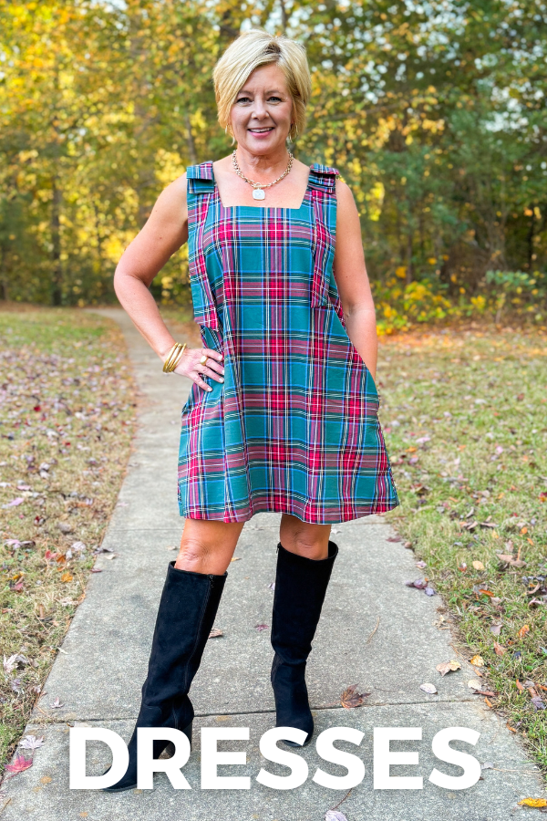 Woman wearing a plaid dress with knee-high boots on a path with trees in the background, labeled 'DRESSES'.
