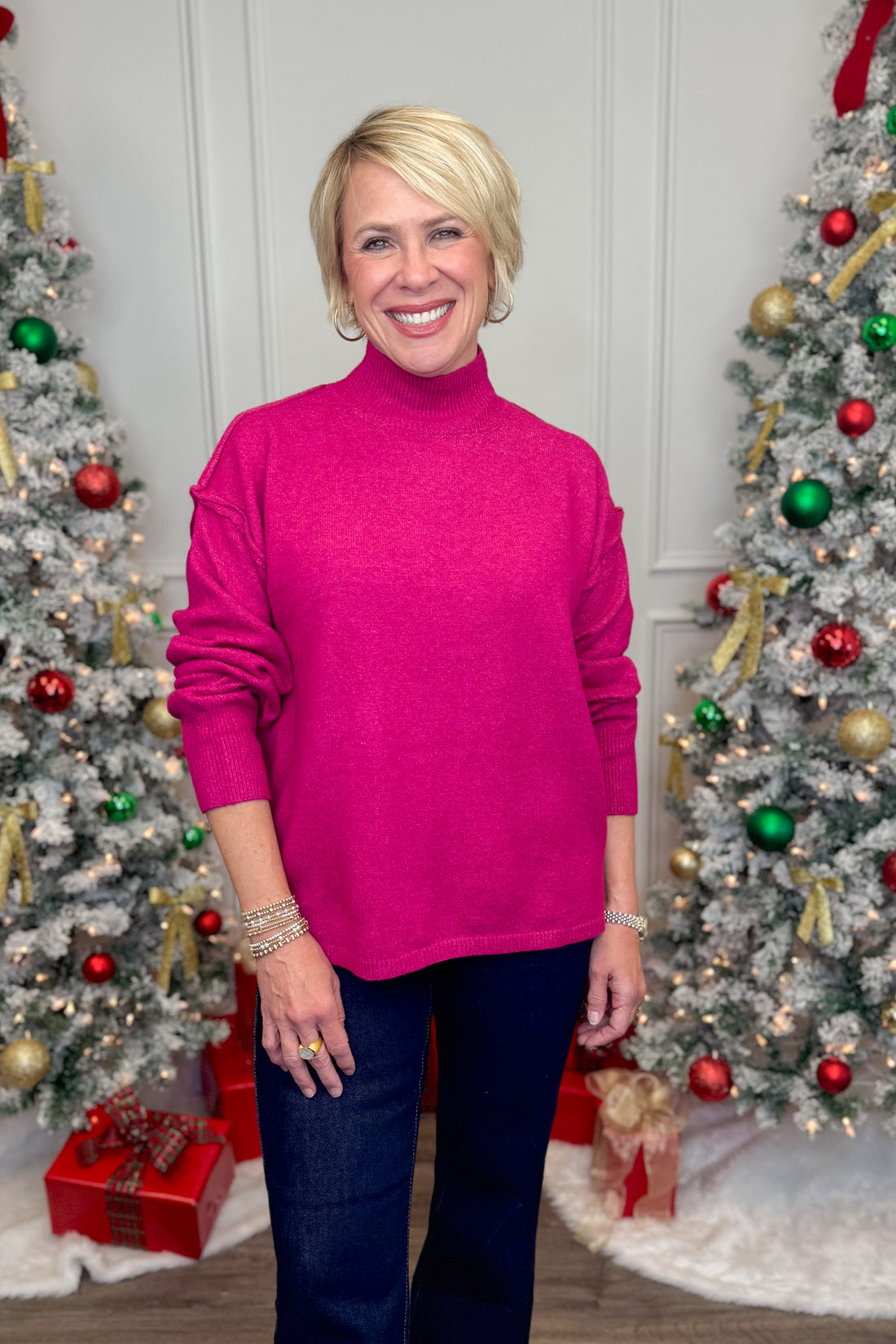 Woman wearing a bright pink sweater standing in front of two decorated Christmas trees.