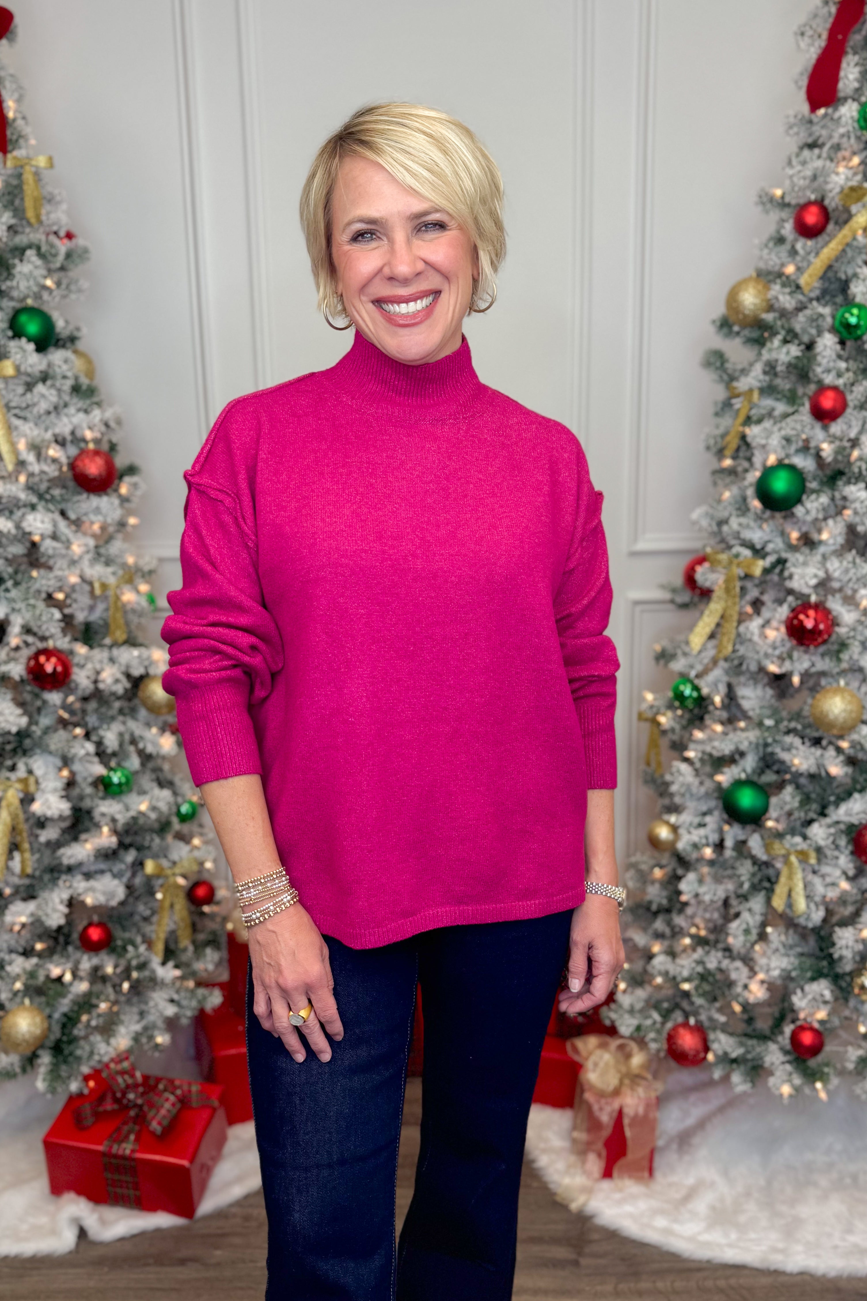 Woman wearing a bright pink sweater standing in front of two decorated Christmas trees.