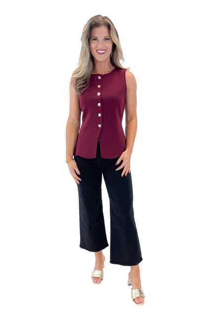 Front view of woman wearing Burgundy Button Detail Sleeveless Peplum Sweater with jeans against white studio background.