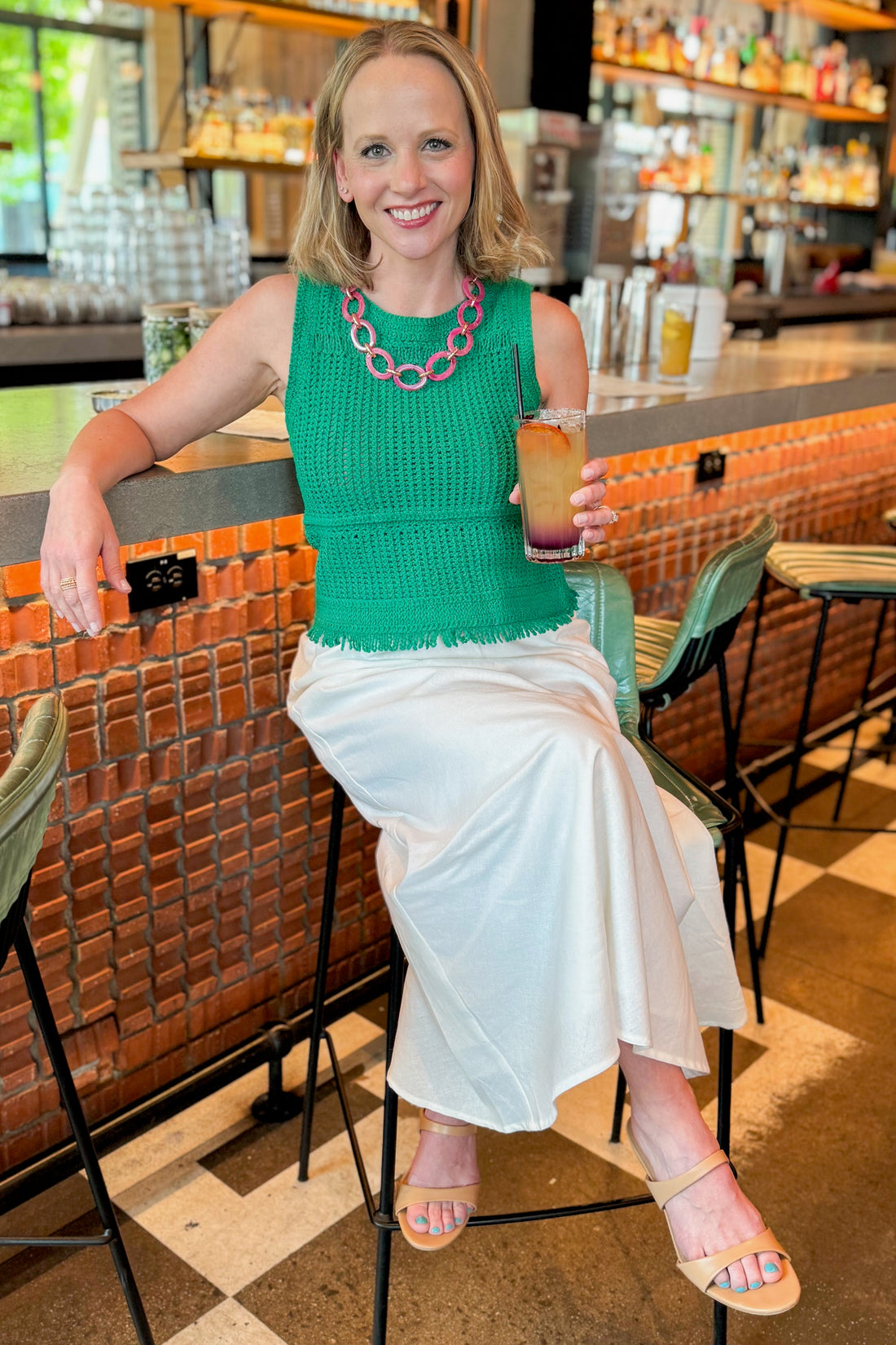 Front view of woman wearing Green Crochet Fringe Detail Tank by Skies Are Blue with white skirt sitting at a bar.