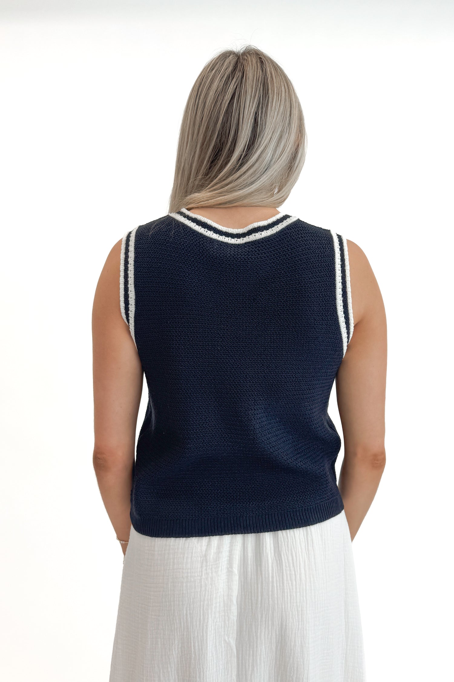 Back view of woman wearing Navy With White Lining Sweater Tank by Skies Are Blue with skirt against white studio background.