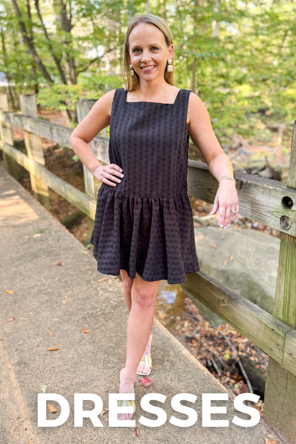Woman wearing a black dress standing outdoors near a wooden fence with 'DRESSES' text overlay.