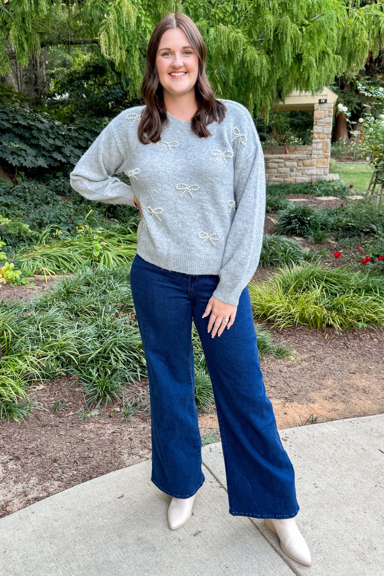 Front view of woman outside with greenery around wearing Grey Pearl Bow Detail Sweater and jeans.  