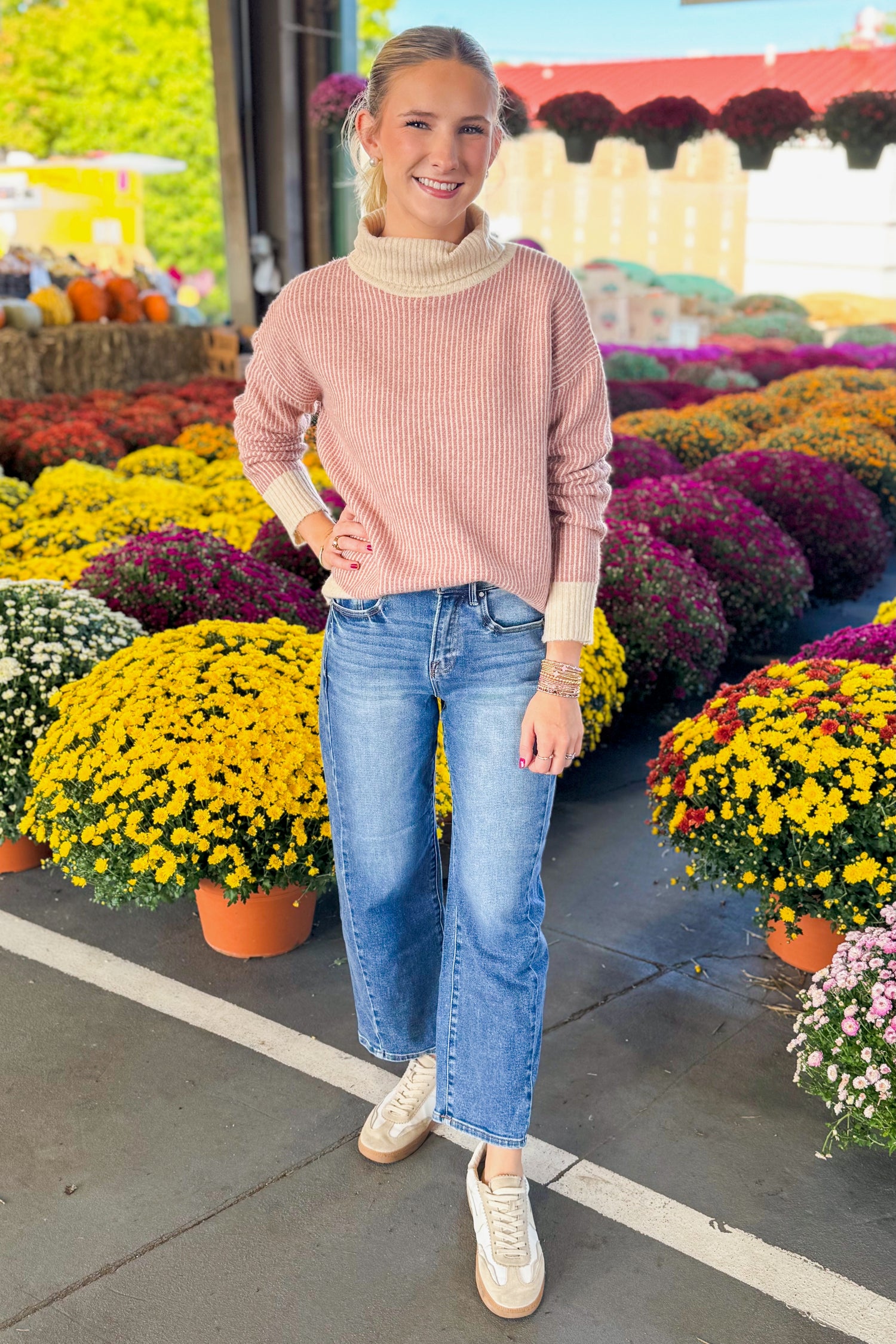 Outside picture of woman wearing Mauve Striped Turtleneck Sweater and blue jeans with mums in the background.