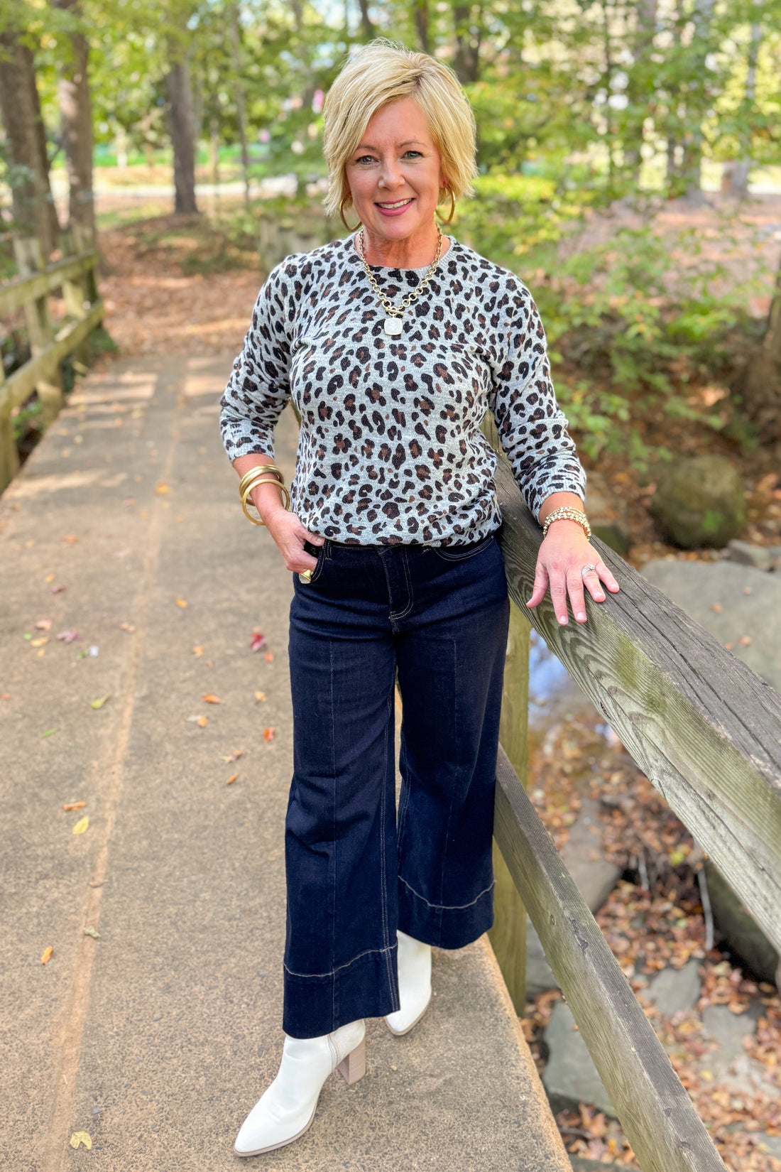 Front view of woman outside wearing Grey Leopard Crewneck Sweater with jeans against with leaves and trees in the background.