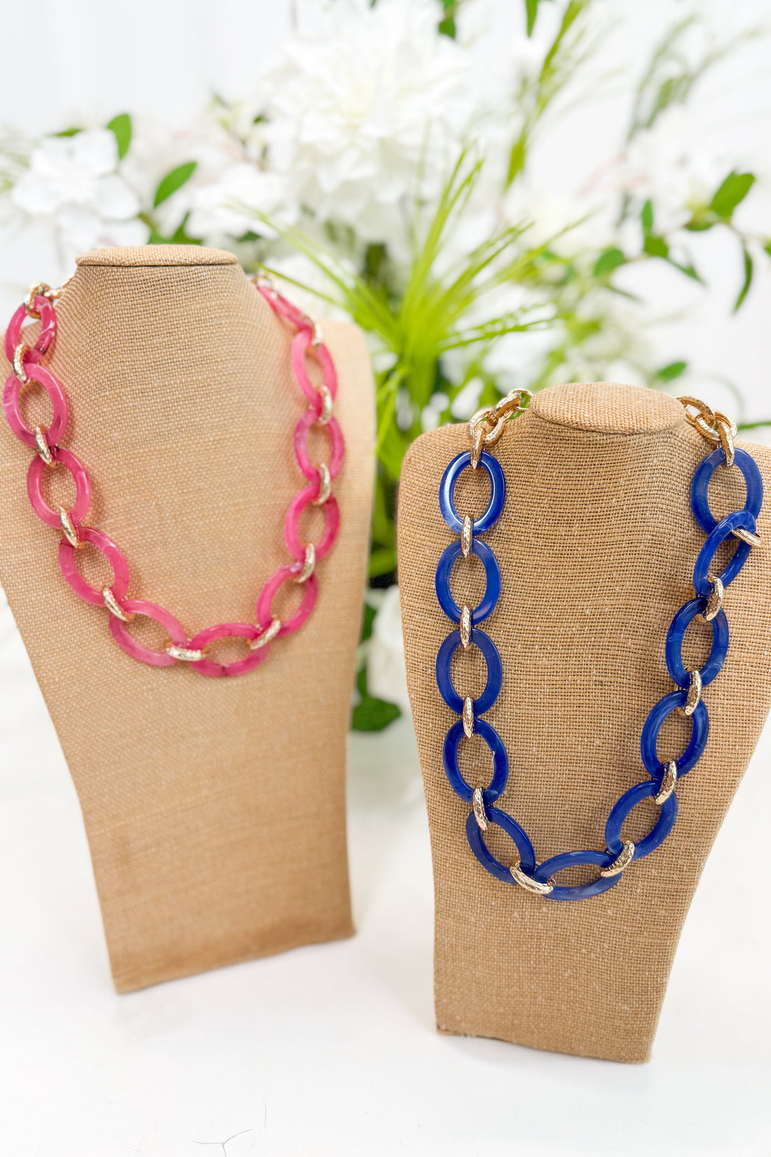 Two necklaces, one pink and one blue, on beige display stands with a blurred floral background.