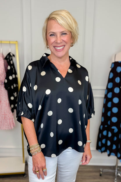 Front view of woman wearing Swagger Boutique’s Black &amp; Cream Polka Dot Satin Top with jeans with clothing hanging in the background.
