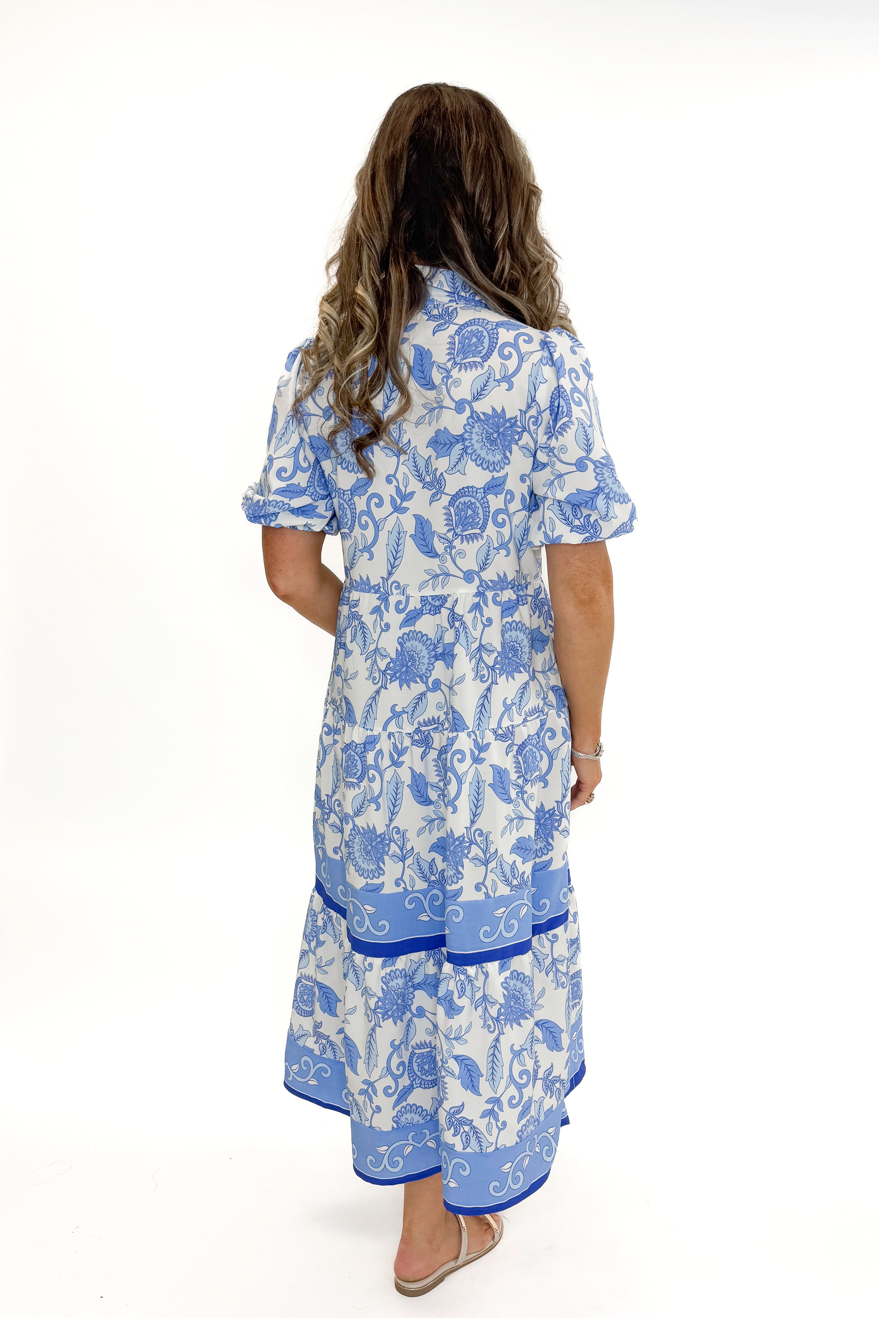Back view of woman wearing Blue Floral Eloise Dress by Mary Square with sandals against white studio background.