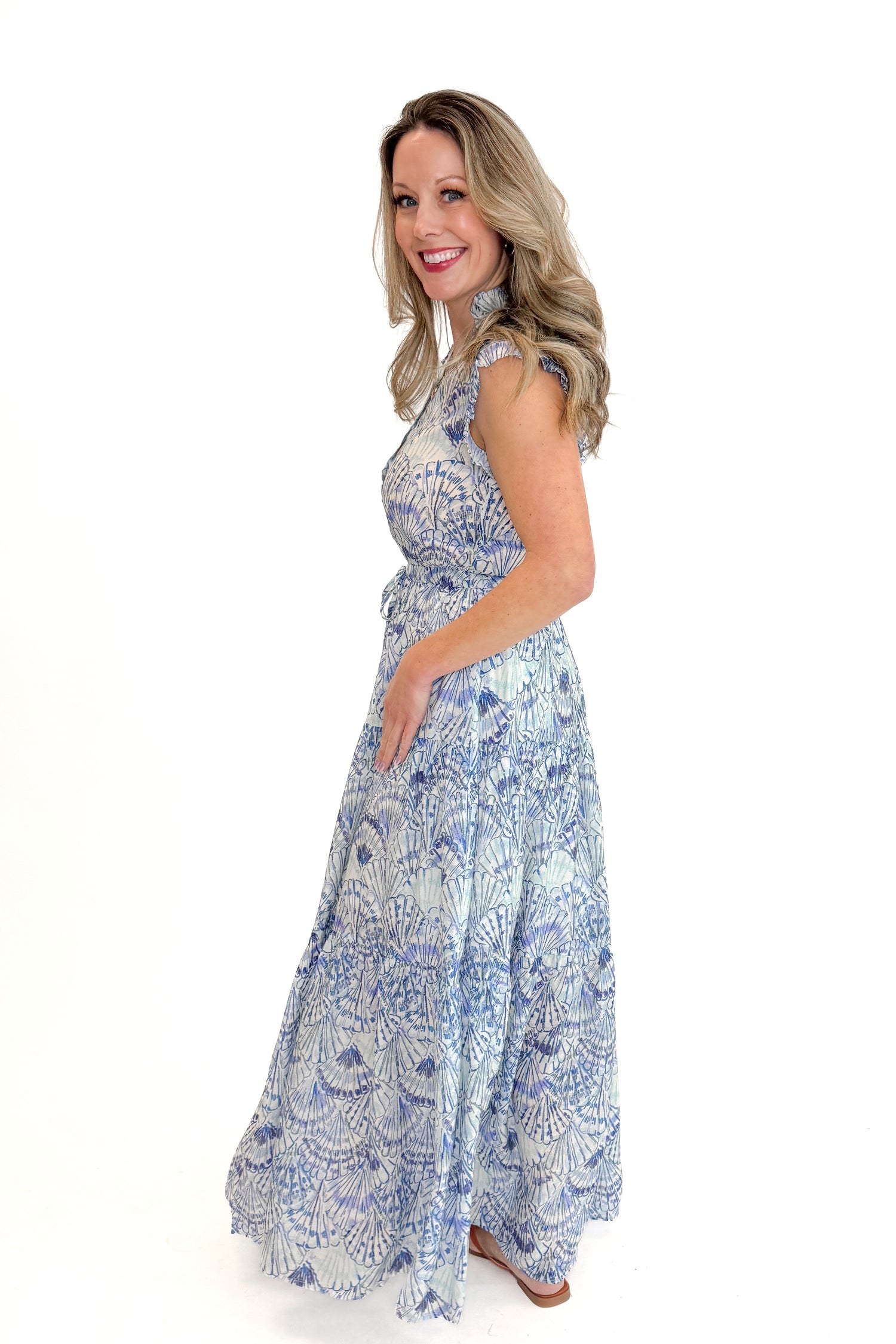 Side view of woman wearing Blue Shell Print Tiered Maxi Dress by PINCH with sandals against white studio background.