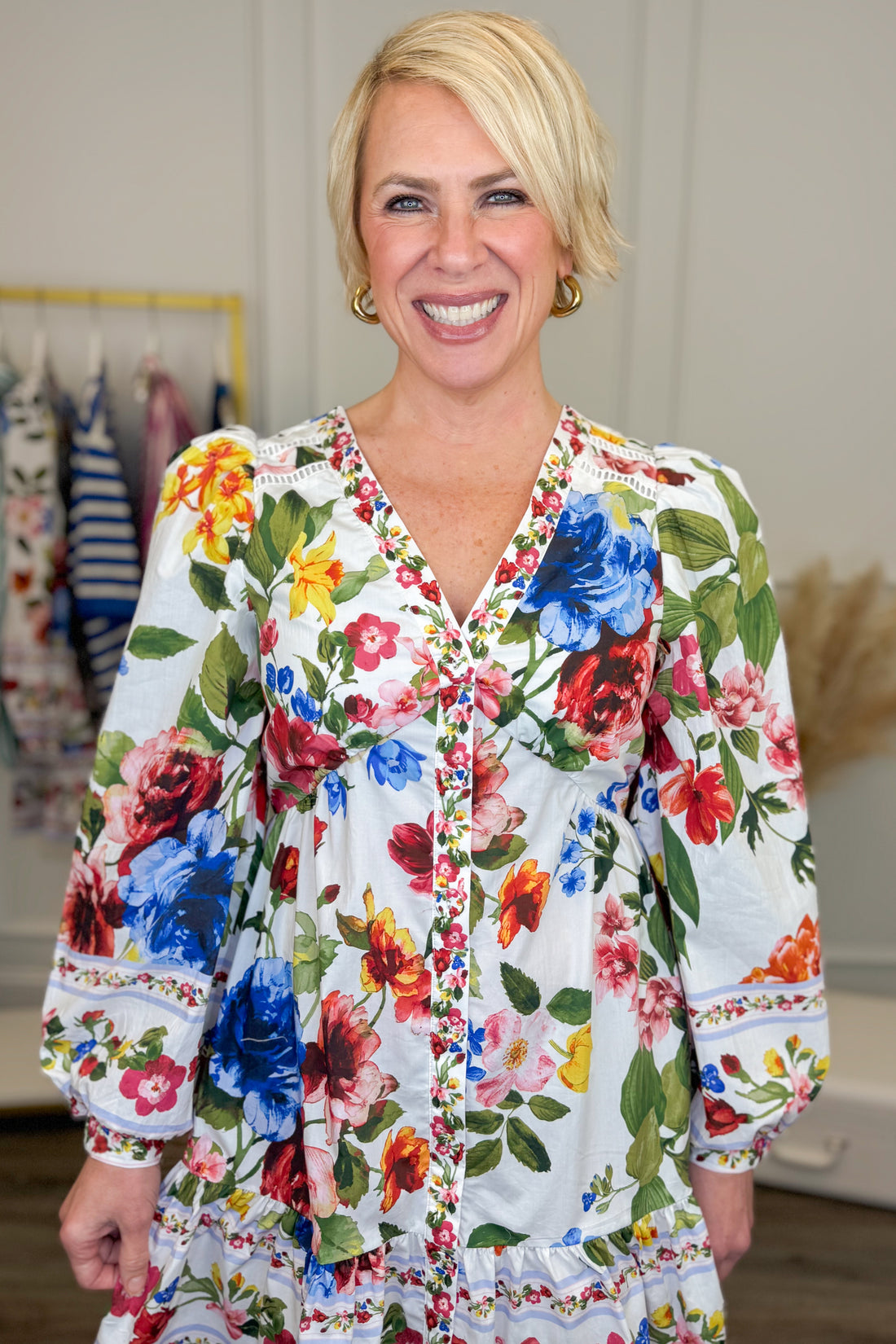 Front detail view of woman wearing Multi Floral Border Print Dress by Skies Are Blue with clothes hanging in the background.
