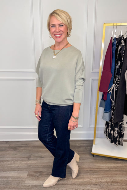 Woman wearing a green sweater and dark jeans standing in front of a clothing rack.