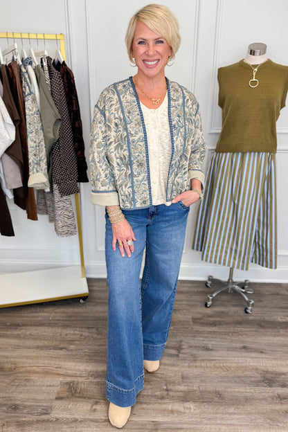 Front view of woman wearing Swagger Boutique’s Beige &amp; Blue Printed Lightweight Jacket, Cream Sleeveless Button up Sweater with Maya Denim Jeans with clothing hanging in the background.