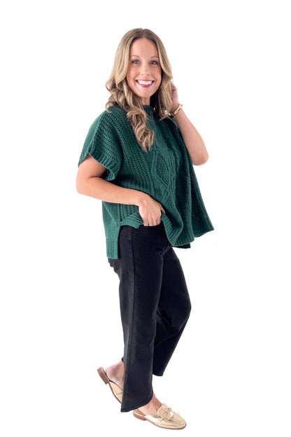 Side view of woman wearing Green Claude Cable Knit Sweater with jeans against white studio background.