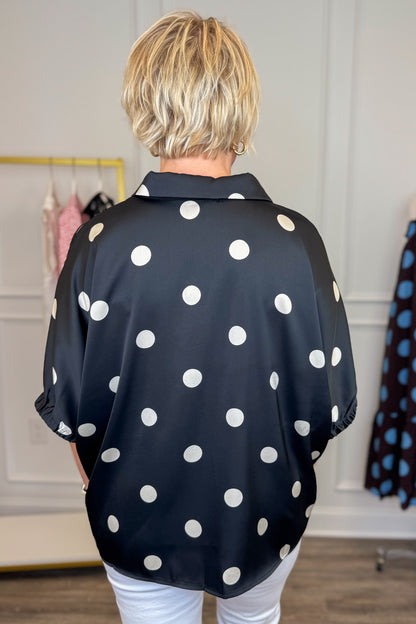 Back view of woman wearing Swagger Boutique’s Black &amp; Cream Polka Dot Satin Top with jeans with clothing hanging in the background.