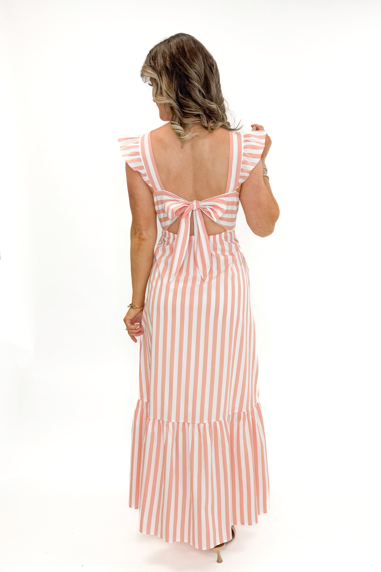 Back view of woman wearing Coral Striped Tie Back Maxi Dress by Skies Are Blue against white studio background.