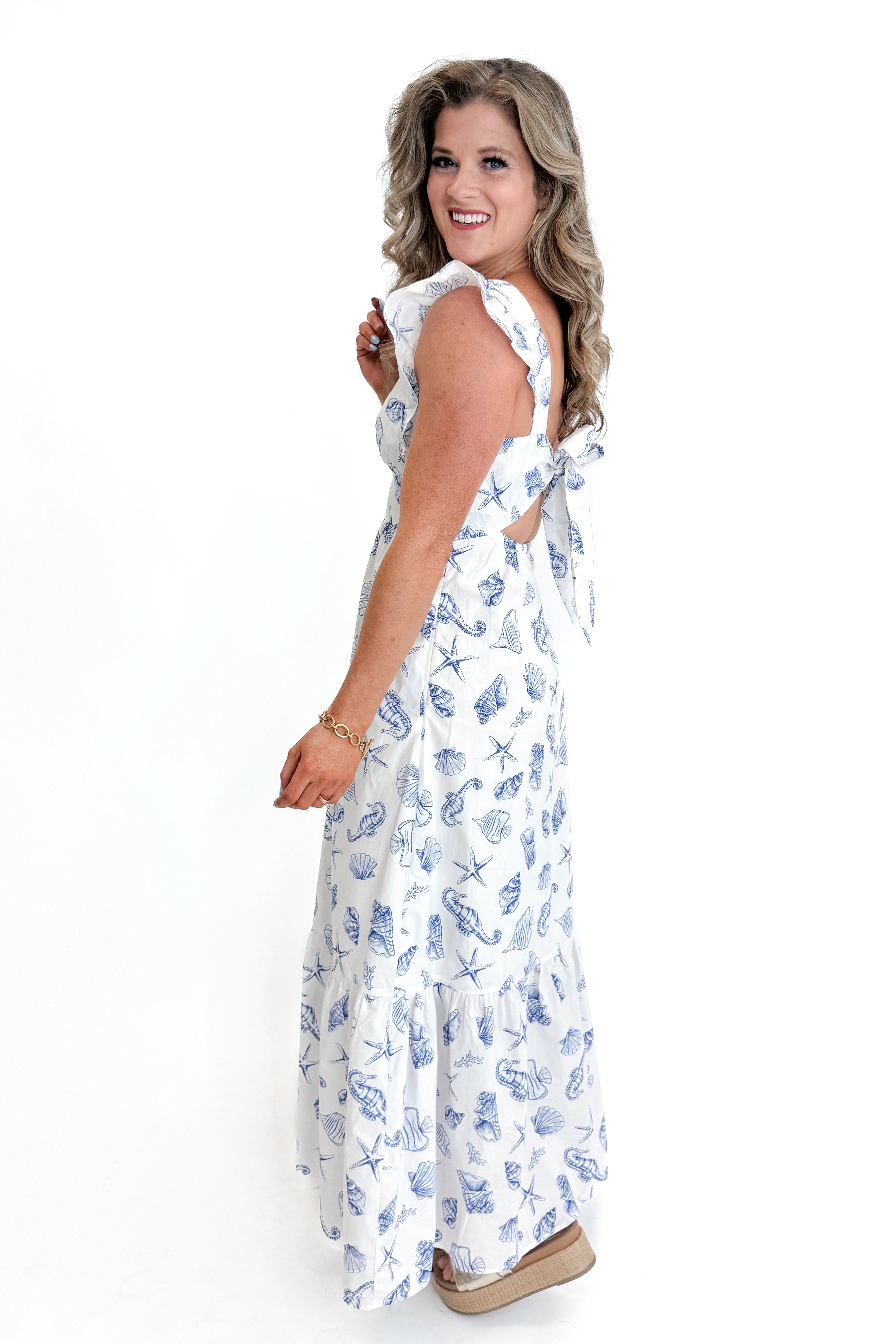 Side view of woman wearing White &amp; Blue Shell Print Tie Back Maxi Dress by Skies Are Blue against white studio background.