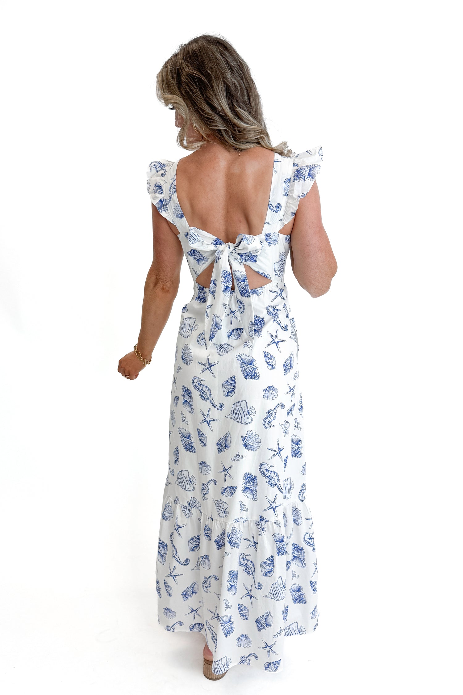 Back view of woman wearing White &amp; Blue Shell Print Tie Back Maxi Dress by Skies Are Blue against white studio background.