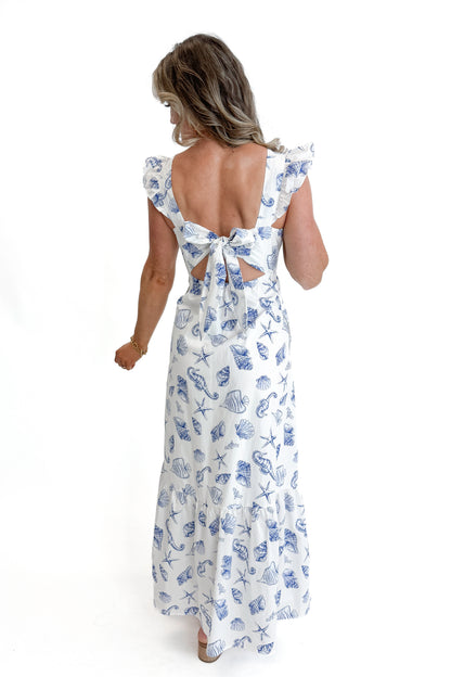 Back view of woman wearing White &amp; Blue Shell Print Tie Back Maxi Dress by Skies Are Blue against white studio background.