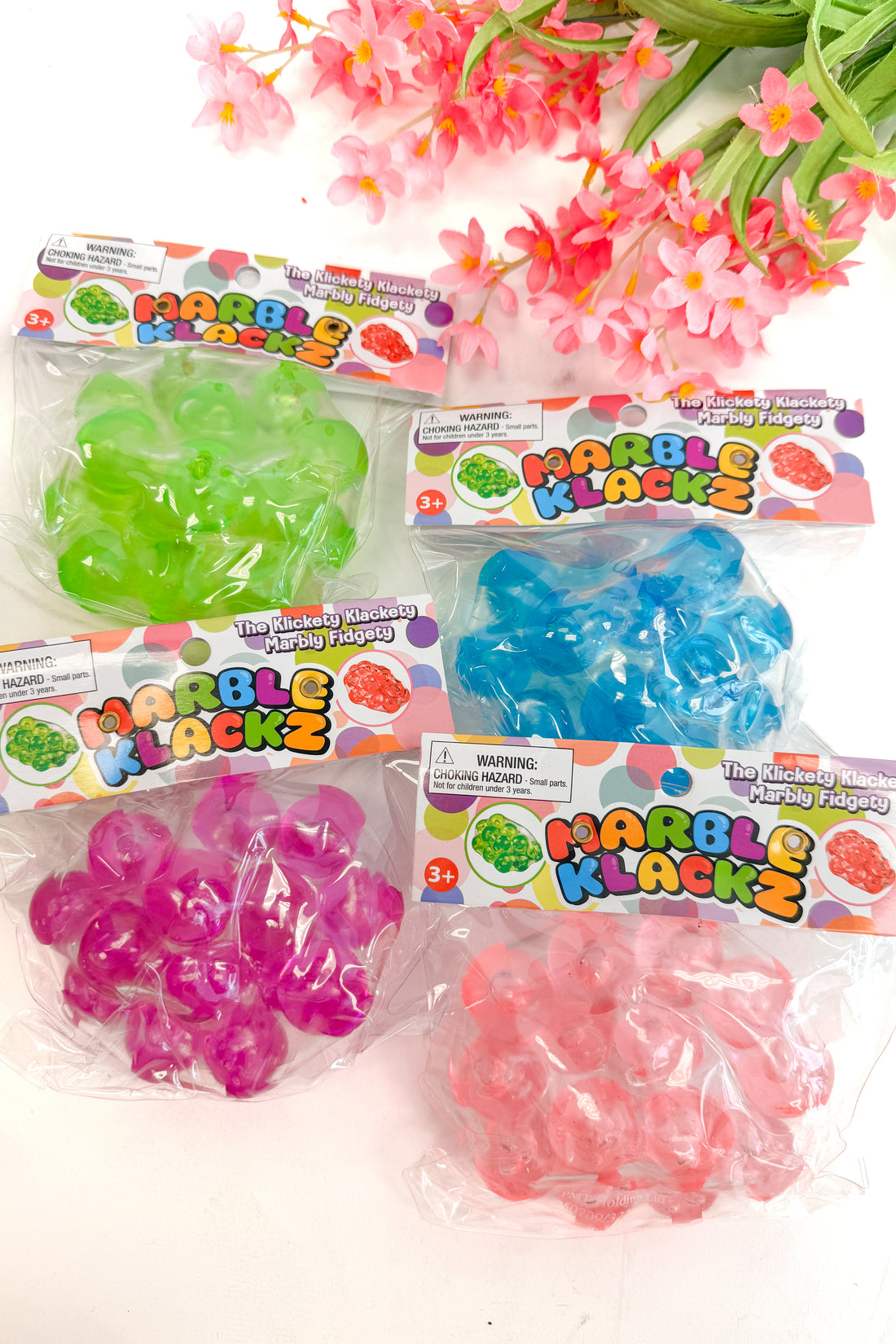 Four packages of Marble Klackz toys in green, blue, pink, and purple on a white background with flowers.