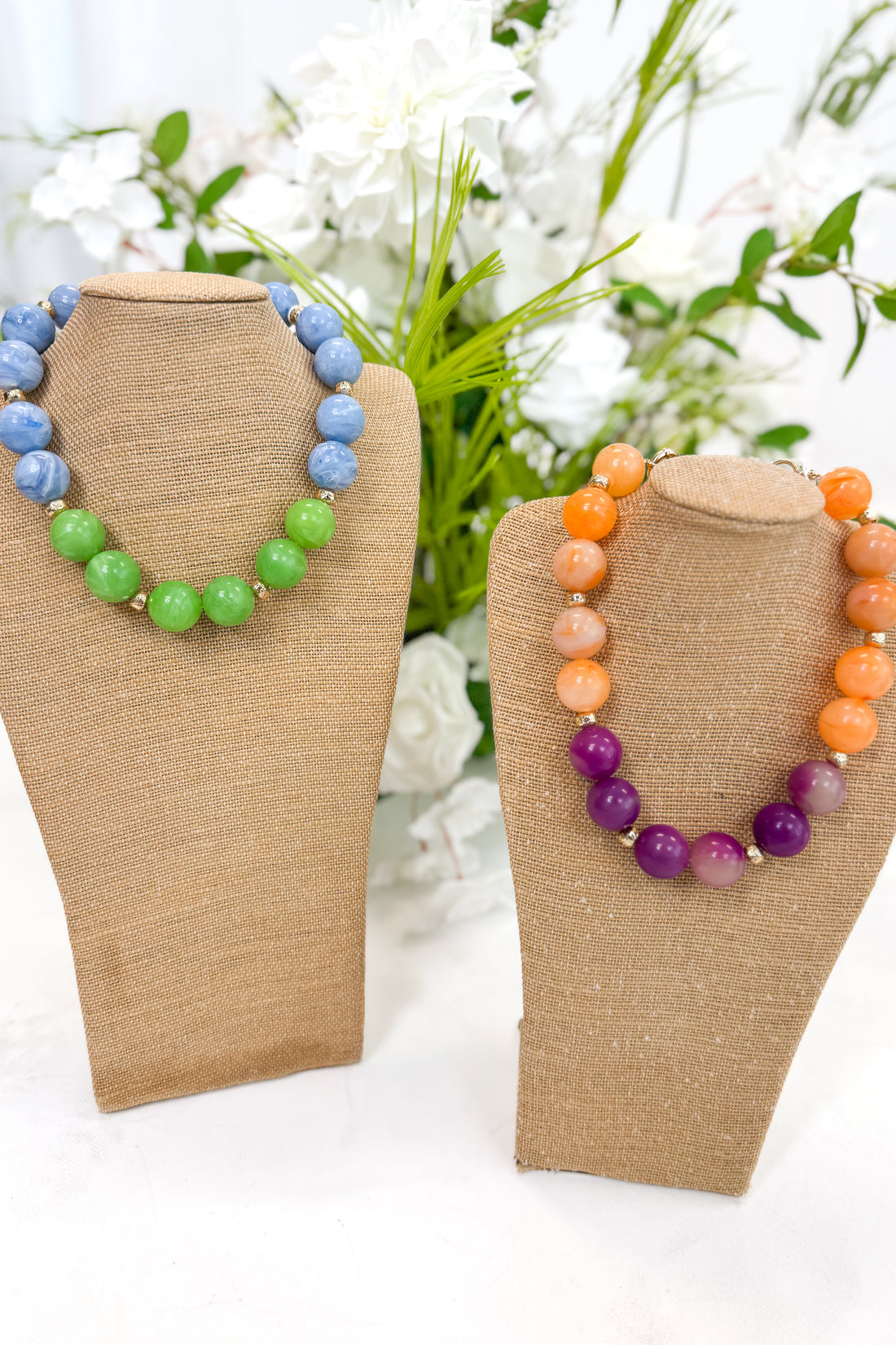 Two colorful beaded necklaces on brown mannequin stands with a floral background.