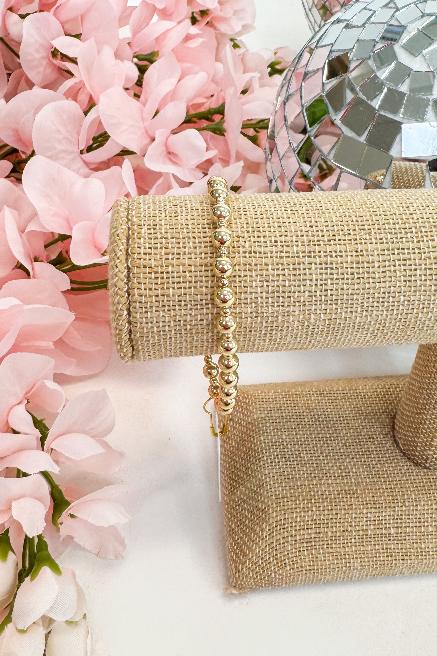 Gold beaded bracelet on a gold jewelry stand with pink flowers in the background