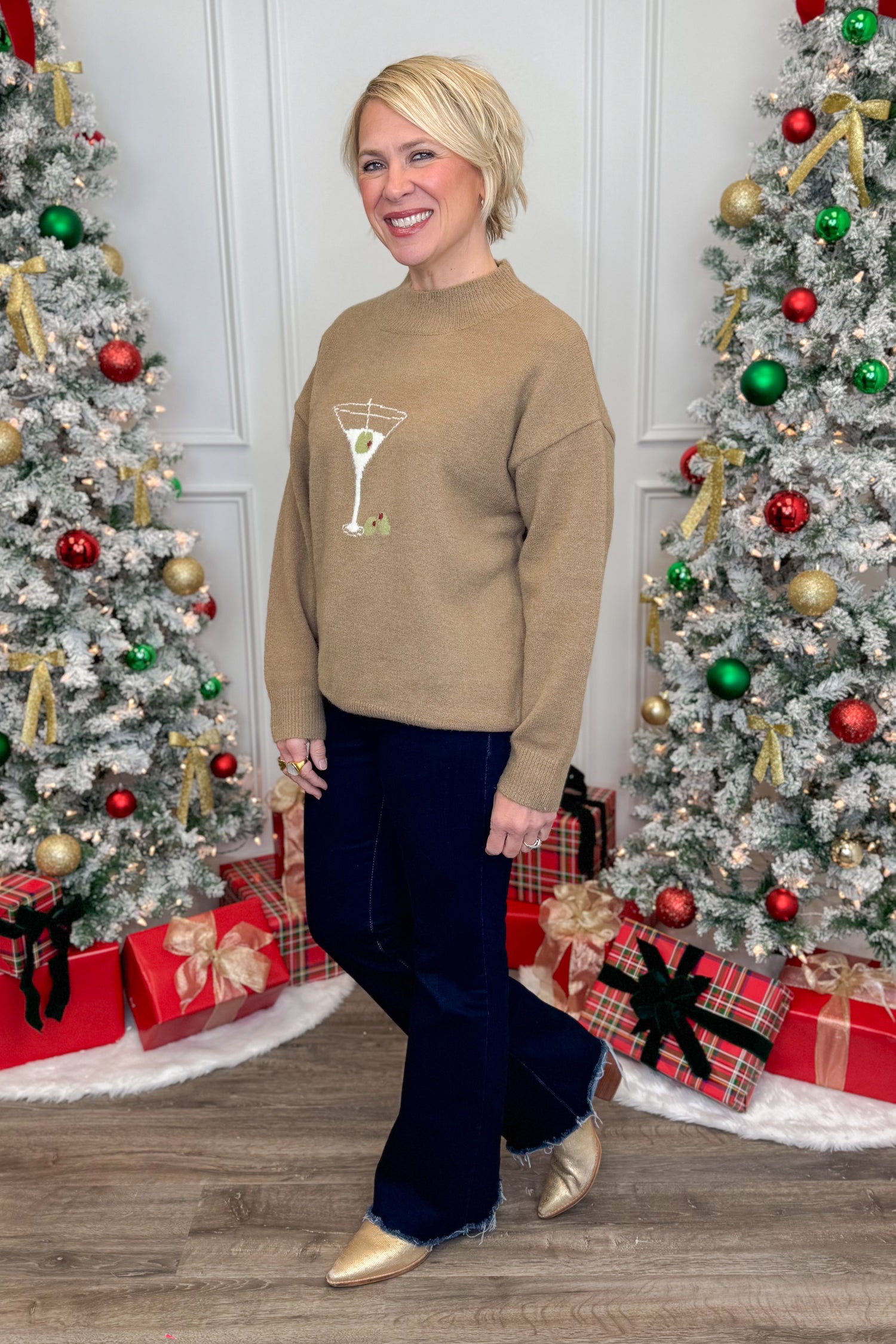 Woman wearing a beige sweater with a martini glass design in front of Christmas trees and presents.