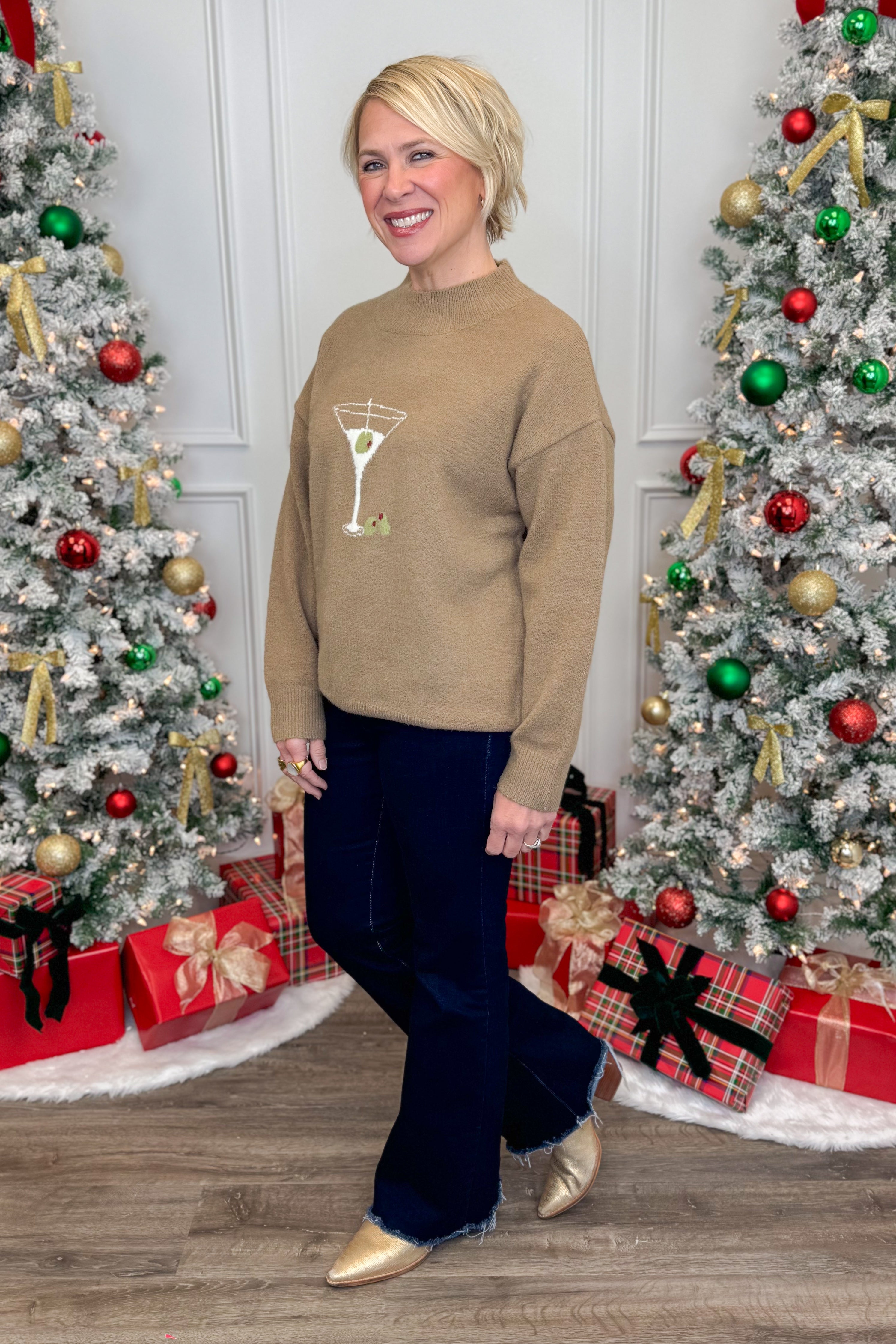 Woman wearing a beige sweater with a martini glass design in front of Christmas trees and presents.