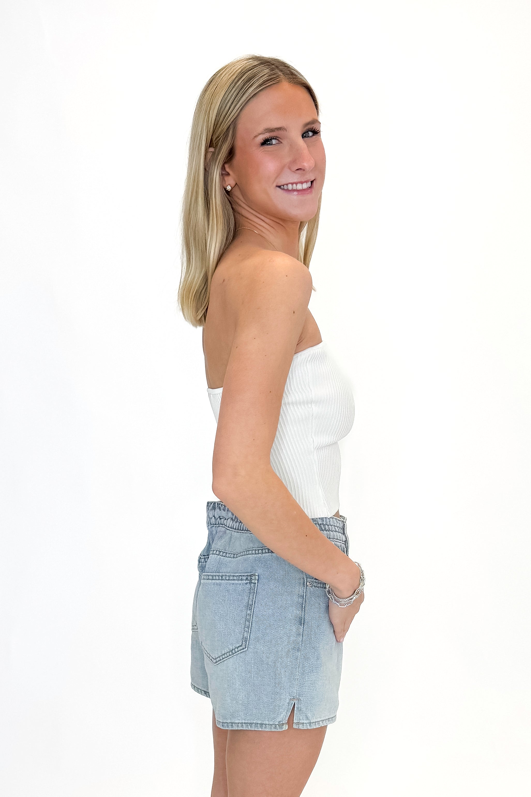 Side view of woman wearing Off White Ribbed Tube Top with jean shorts against white studio background.