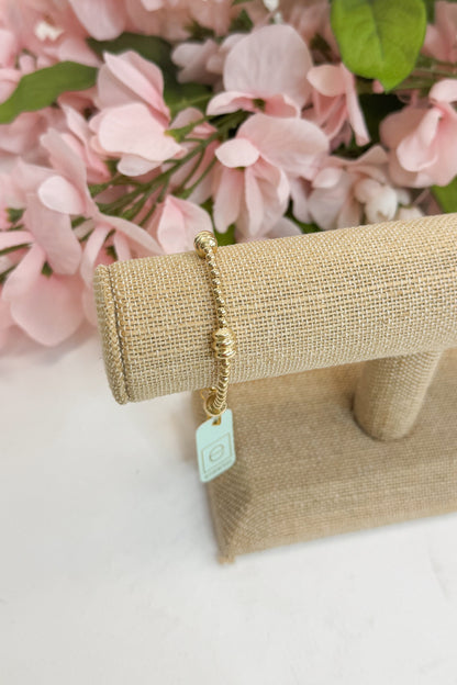 Gold bracelet with a tag on a beige stand against a pink floral background