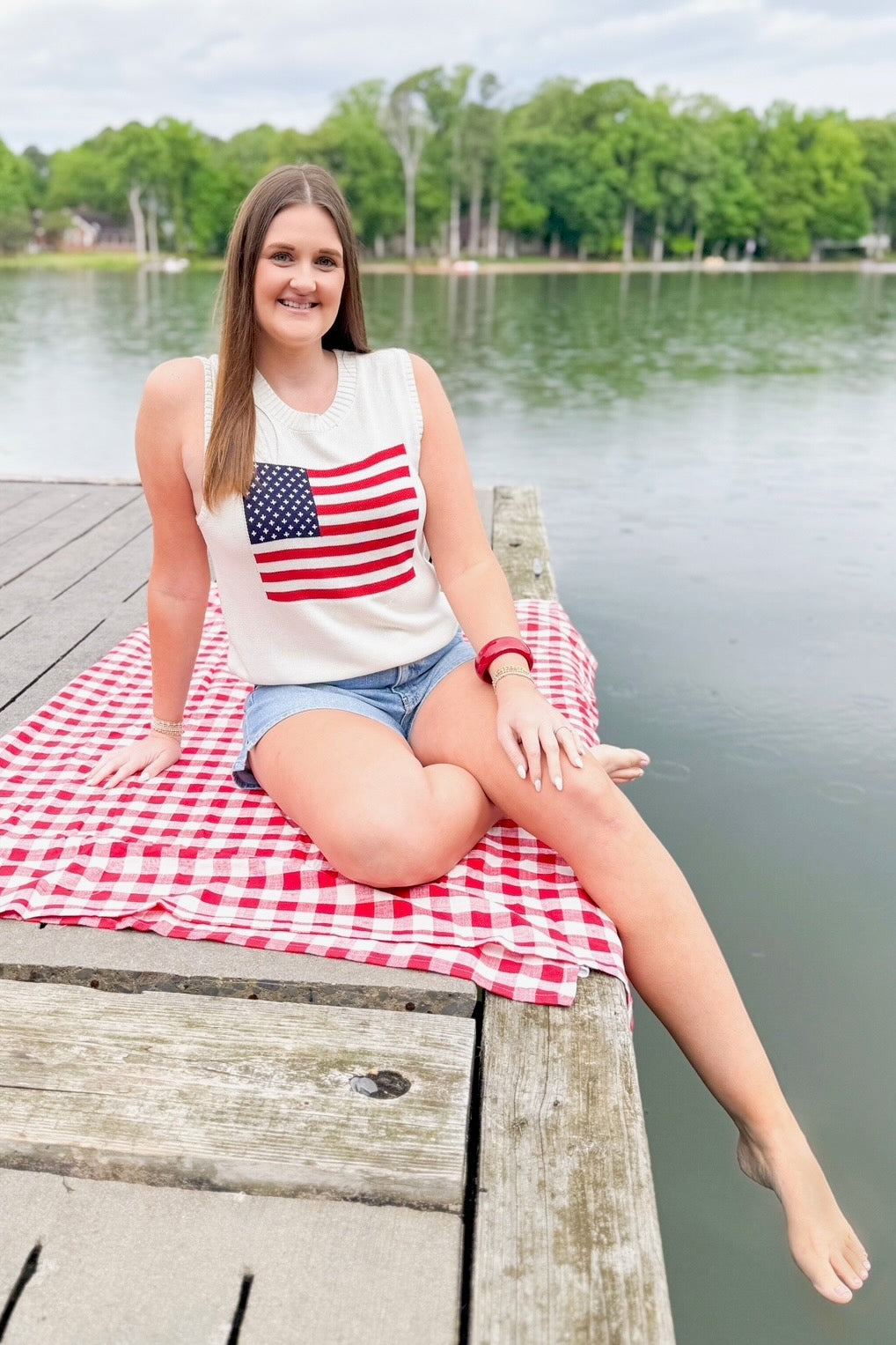 Front view of woman wearing Swagger Boutique’s American Flag Sweater Tank with jean shorts sitting outside at a lake.