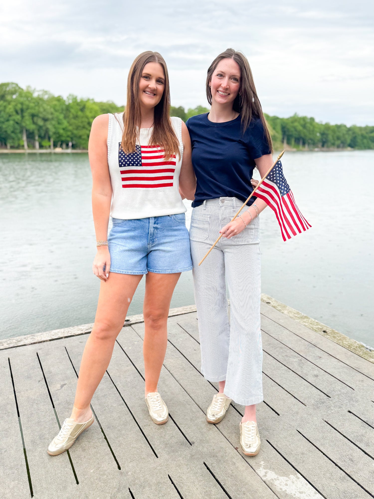 Front view of woman wearing Swagger Boutique’s American Flag Sweater Tank with jean shorts and woman wearing Z Supply blue top and Cream & Blue Striped Jeans With Patch Pocket by Just Black Denim outside at the lake.