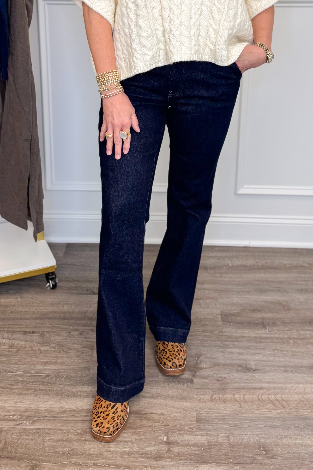 A person wearing dark blue flare jeans, a cream cable-knit top, and leopard-print slip-on shoes, standing on a wood floor in front of a white paneled wall.