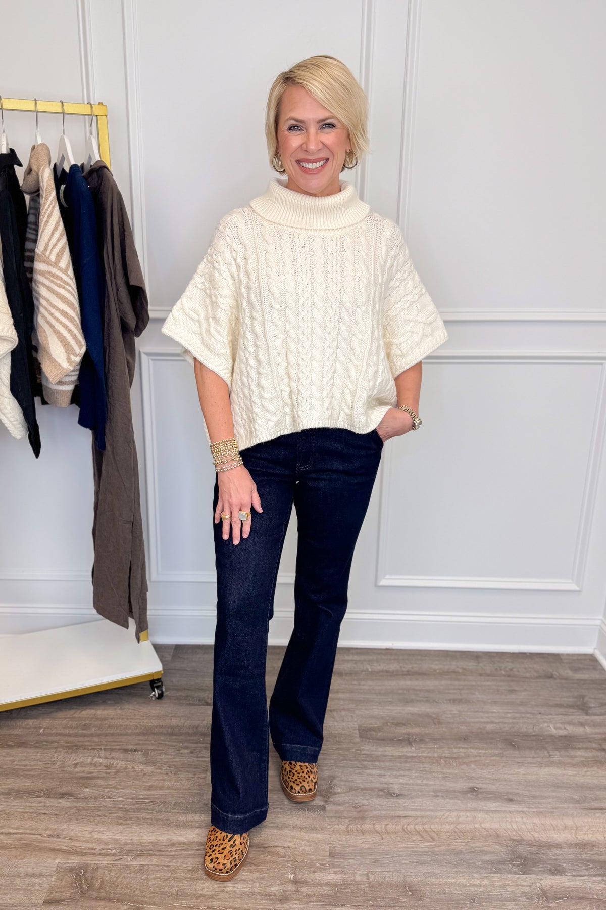 A woman modeling a cream cable-knit turtleneck poncho sweater, paired with dark blue jeans and leopard print shoes, standing in a room with a white paneled wall and a clothing rack.