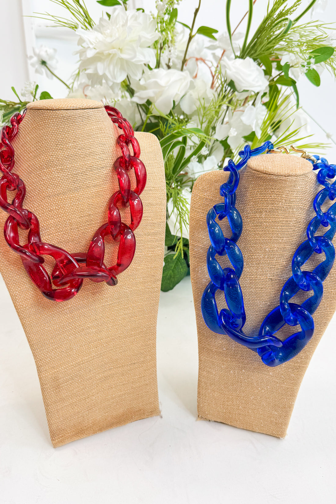 Red and blue chain necklaces on beige display stands with a floral background.
