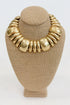 Front view of a gold collar necklace on a beige jewelry bust, featuring chunky curved links arranged in a graduated pattern.