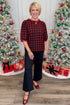 Woman in a red and black patterned top and black pants standing in front of Christmas trees with decorations.