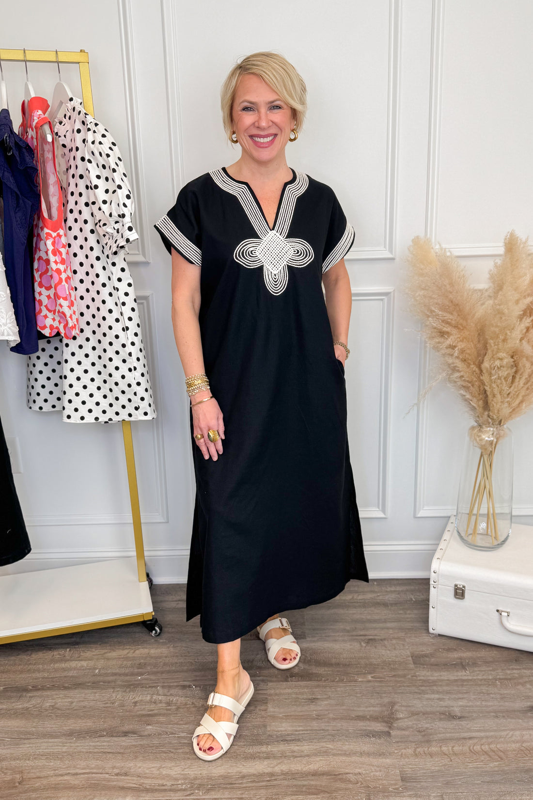 Woman wearing a black dress with white embroidery in a room with a clothing rack and decorative elements.