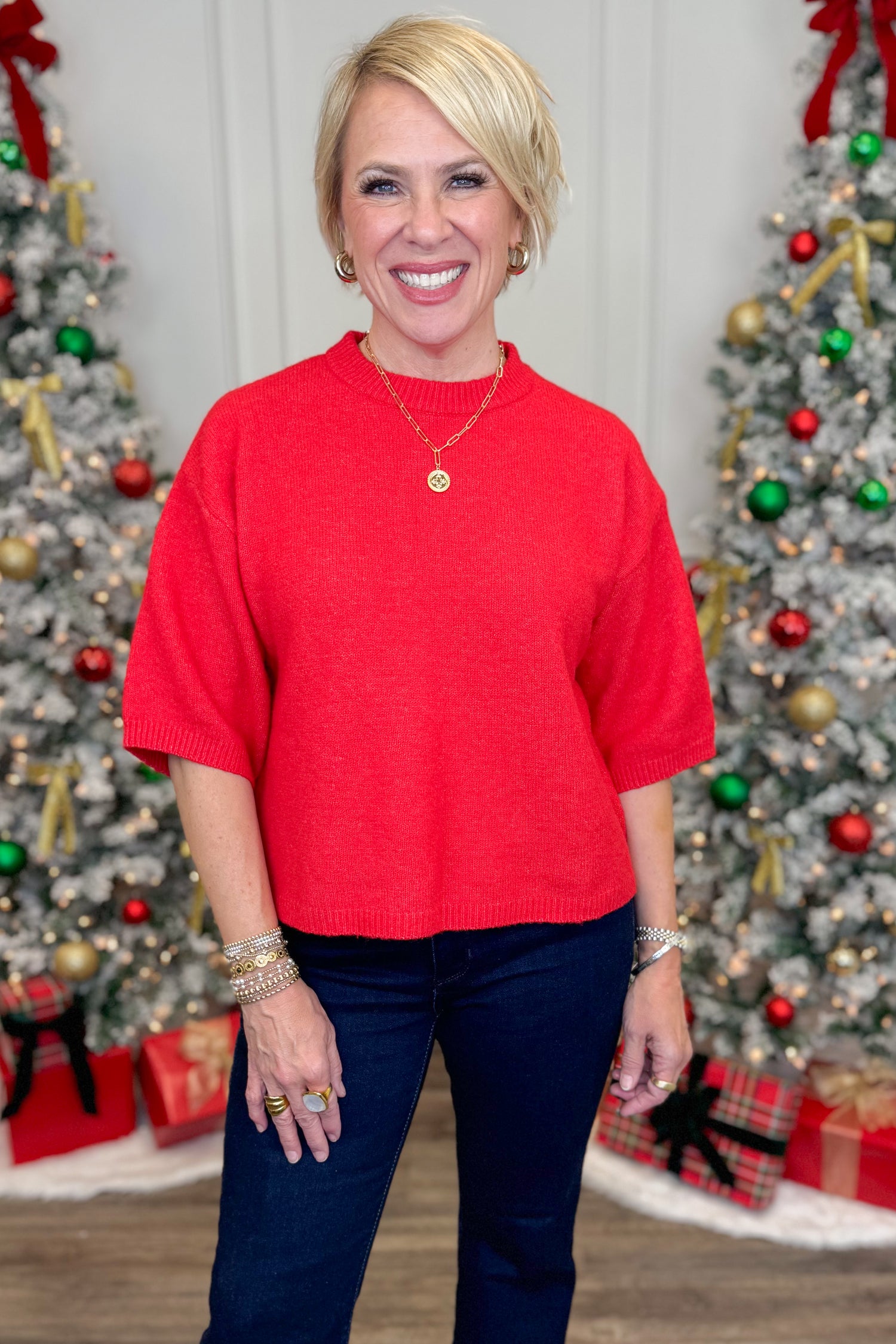 Woman in a red sweater and blue pants standing in front of Christmas trees with decorations.