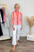Woman wearing a colorful sleeveless top and white pants in a room with a clothing rack and decorative elements.
