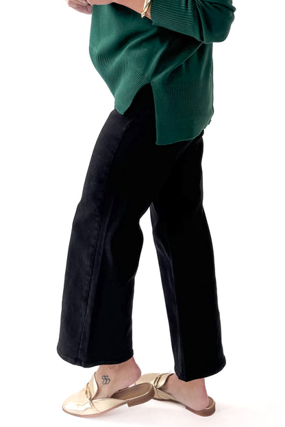 Side view of woman wearing green top with black wide leg jeans against white studio background.