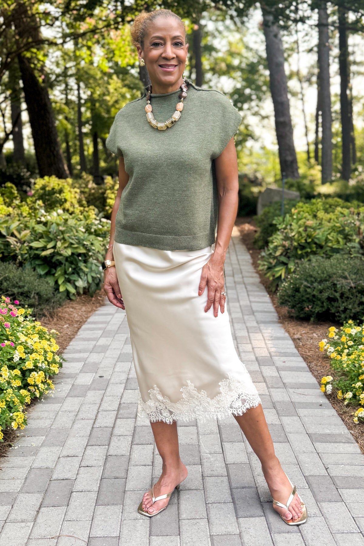 Front view of woman wearing Swagger Boutique’s Beige Satin &amp; Lace Midi Skirt  standing next to a garden outside