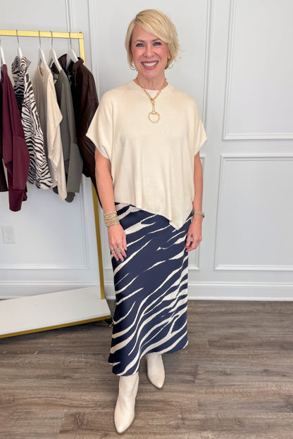 A woman stands indoors modeling a cream short-sleeve top and a navy midi skirt with a white zebra-like stripe pattern. She is wearing cream ankle boots and gold jewelry, with a clothing rack and paneled wall in the background.