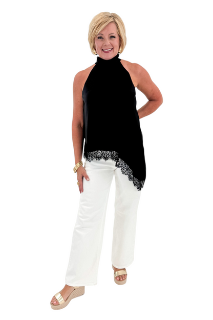 Front view of woman wearing Swagger Boutique’s Black Asymmetrical Halter Neck Satin Lace Top with jeans against white studio background.