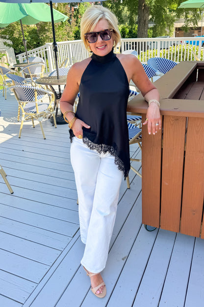 Front view of woman wearing Swagger Boutique’s Black Asymmetrical Halter Neck Satin Lace Top with jeans standing outside at a restaurant.