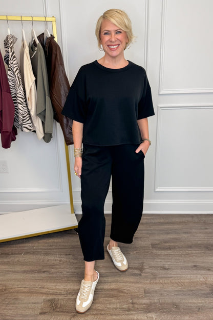 Front view of woman wearing Swagger Boutique’s Black Round Neck Short Sleeve Top with matching barrel pants with clothes hanging in the background.